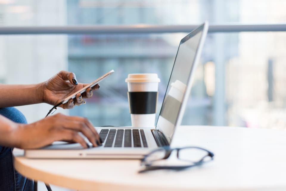 Person using a phone beside a laptop and a cup of coffee